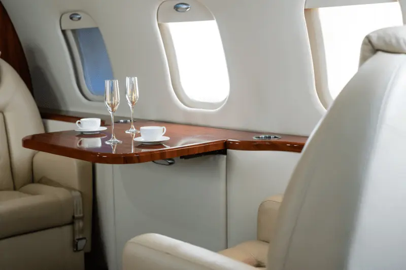 The spacious cabin of a private jet, featuring plush leather seats, a wooden table, and large windows.